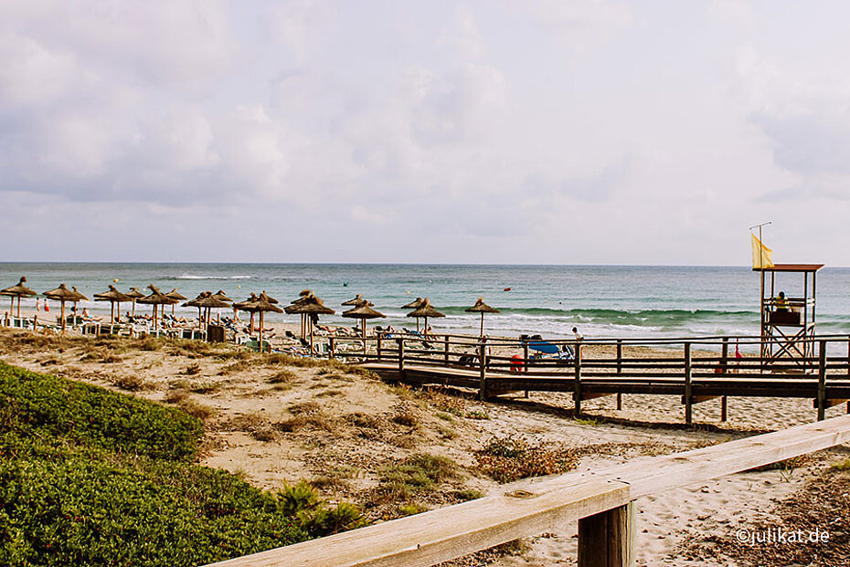 Ein Holzsteg führt hinunter zum breiten Sandstrand mit seinen Strohschirmen.