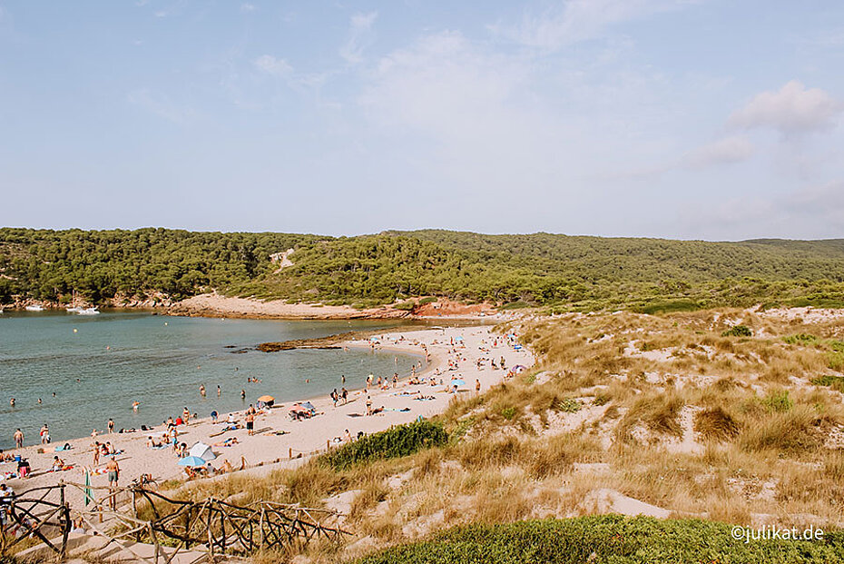 Ein breiter Sandstrand liegt vor einer wilden Dünenlandschaft und roten Felsen.